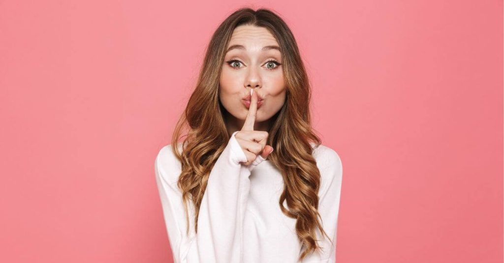 pink background and woman holding finger to mouth implying "shh"